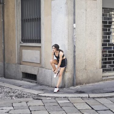 Slim female runner in activewear standing on corner of street and tying shoelaces on sneakers while leaning on stone wall of building and looking away